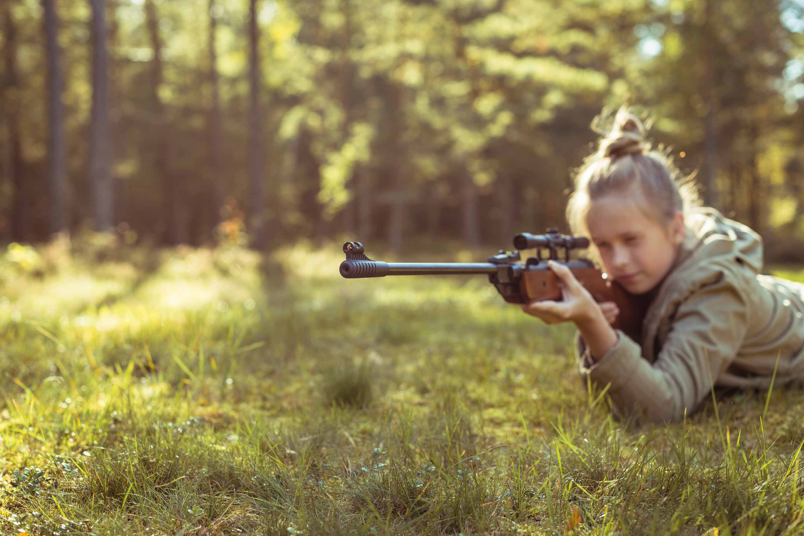 A person lying prone on the grass in a forest, aiming a rifle with a scope.