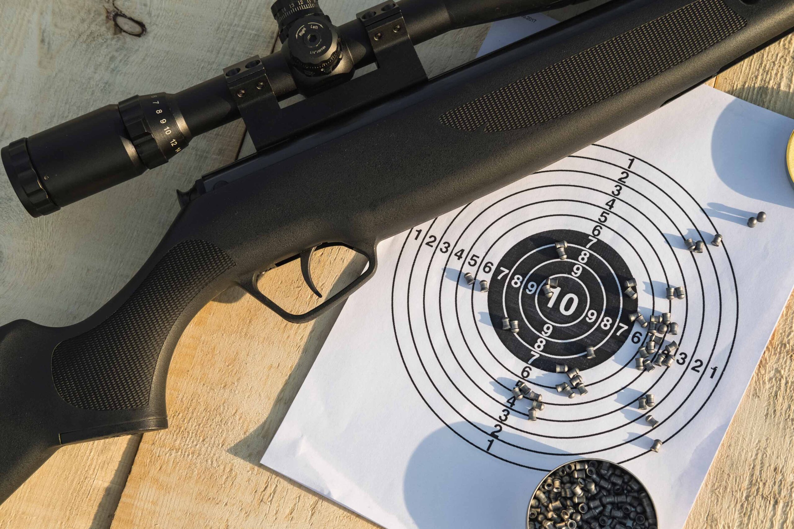 A black air rifle with a mounted scope is placed on a wooden surface next to a paper target. The target has multiple bullet holes, primarily clustered around the center, indicating successful shots. Scattered pellets are visible on and around the target.