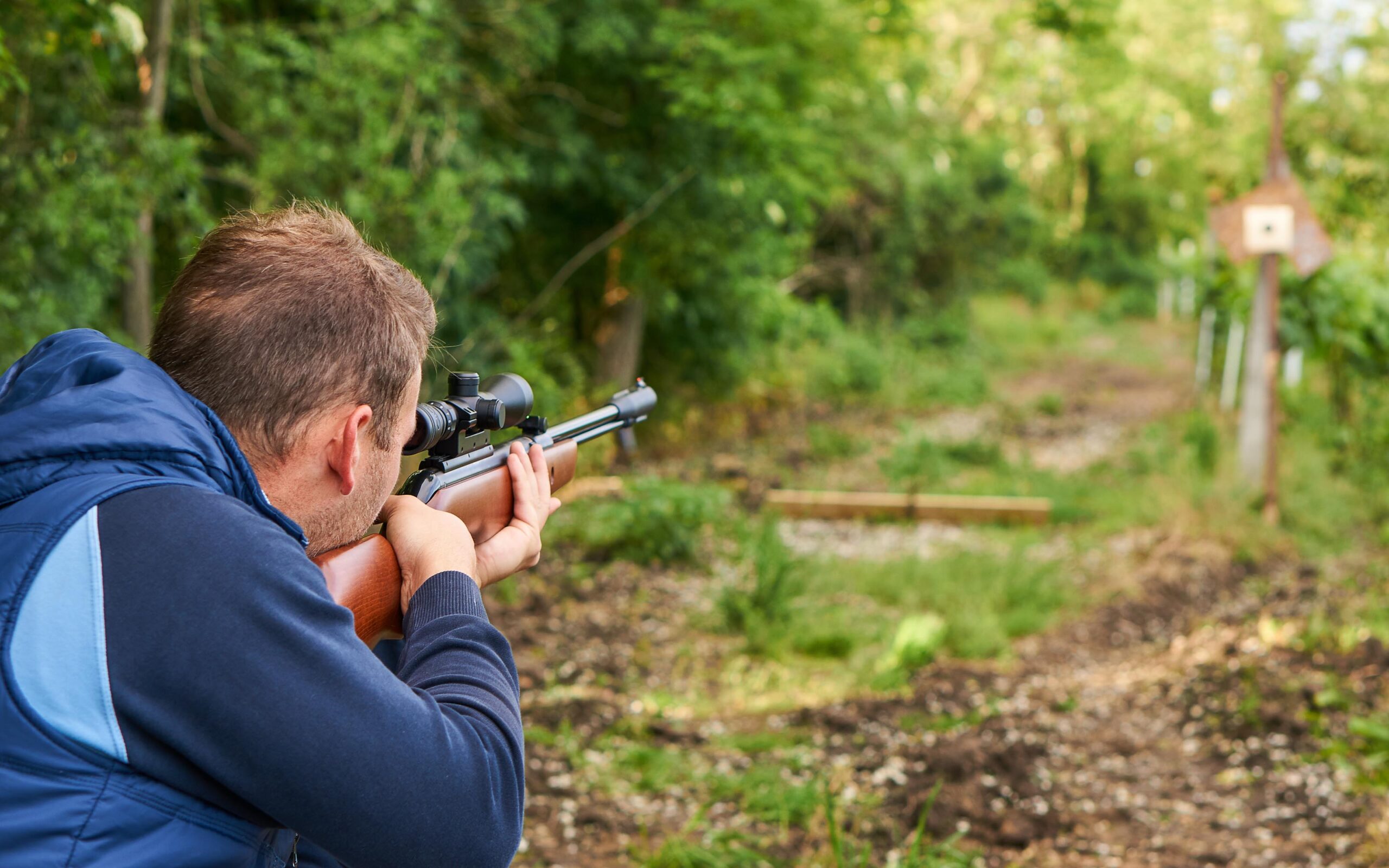 A person in a blue jacket aims a rifle at a target in the distance, set up along a wooded path.