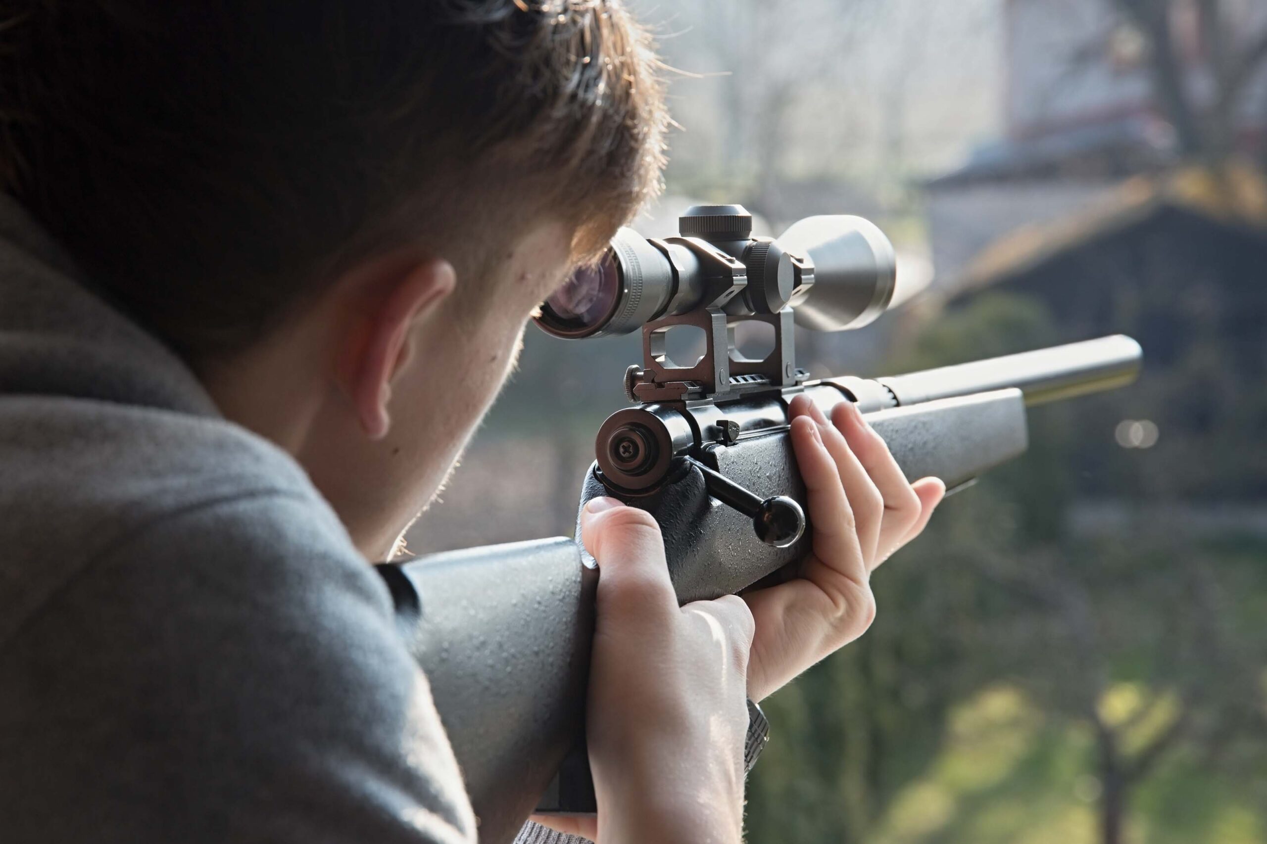 Person aiming a rifle with a scope in an outdoor setting