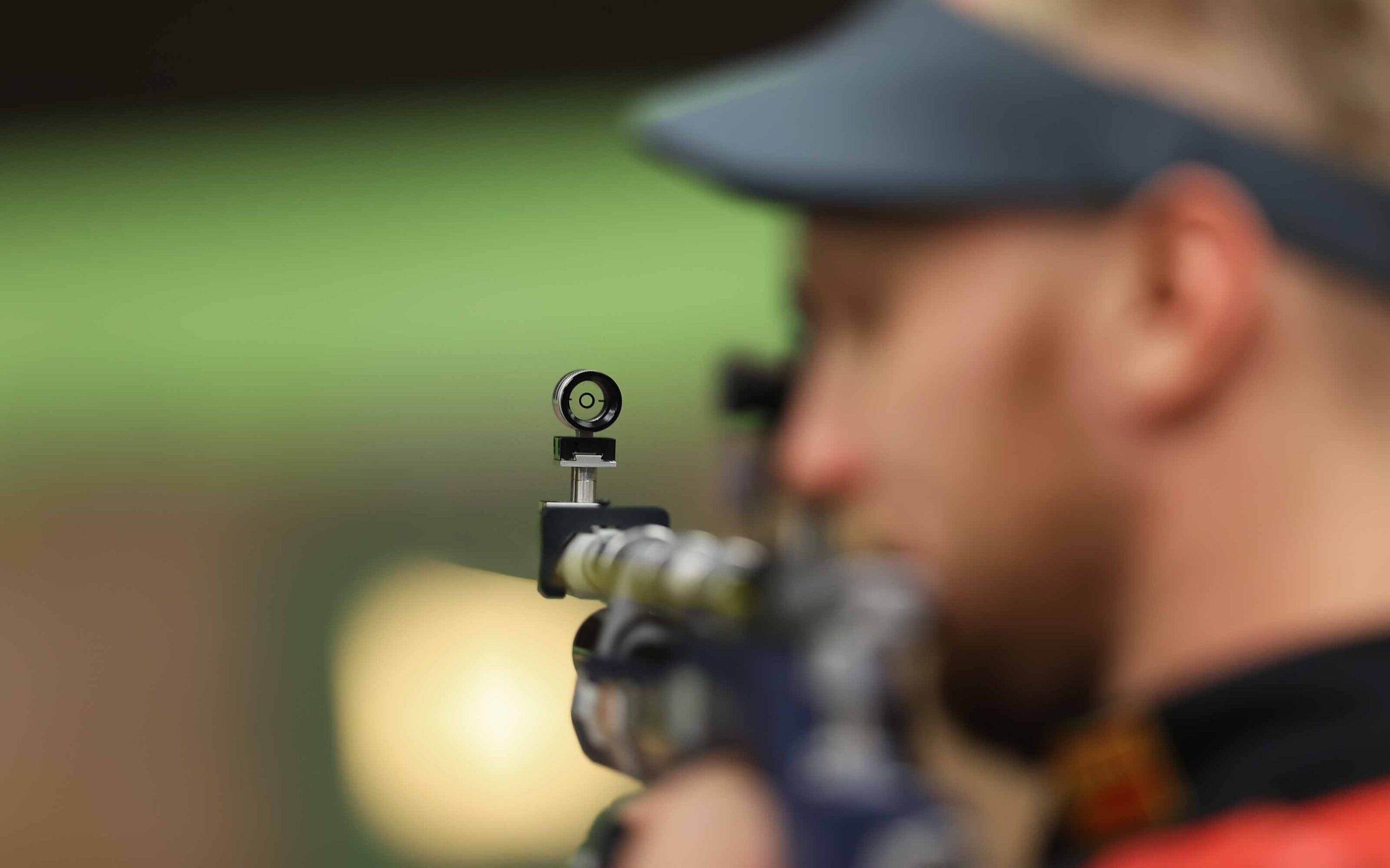 Person aiming a rifle with focus on the sight, wearing a visor; blurred background with green and yellow tones