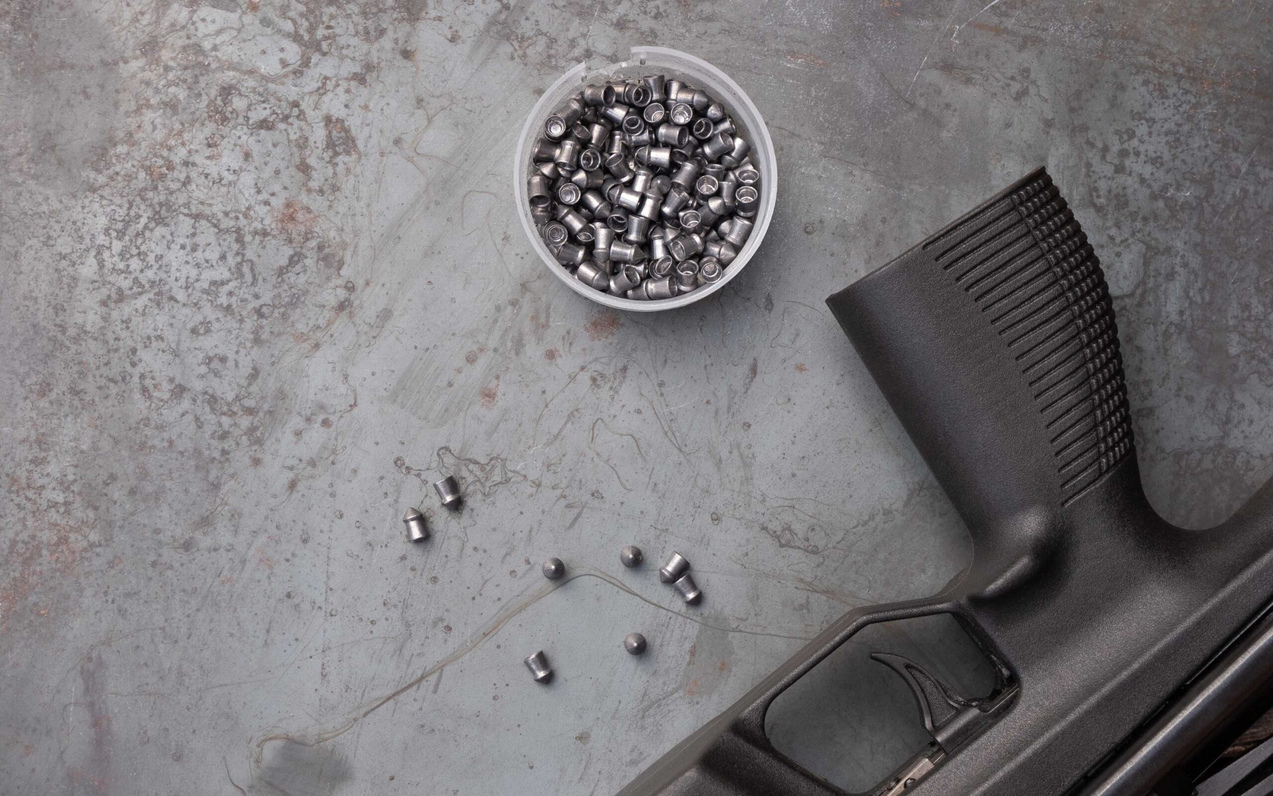 Close-up of a black air rifle grip beside a container of silver airgun pellets on a textured gray surface, illustrating components for airgun sports during National Shooting Sports Month.