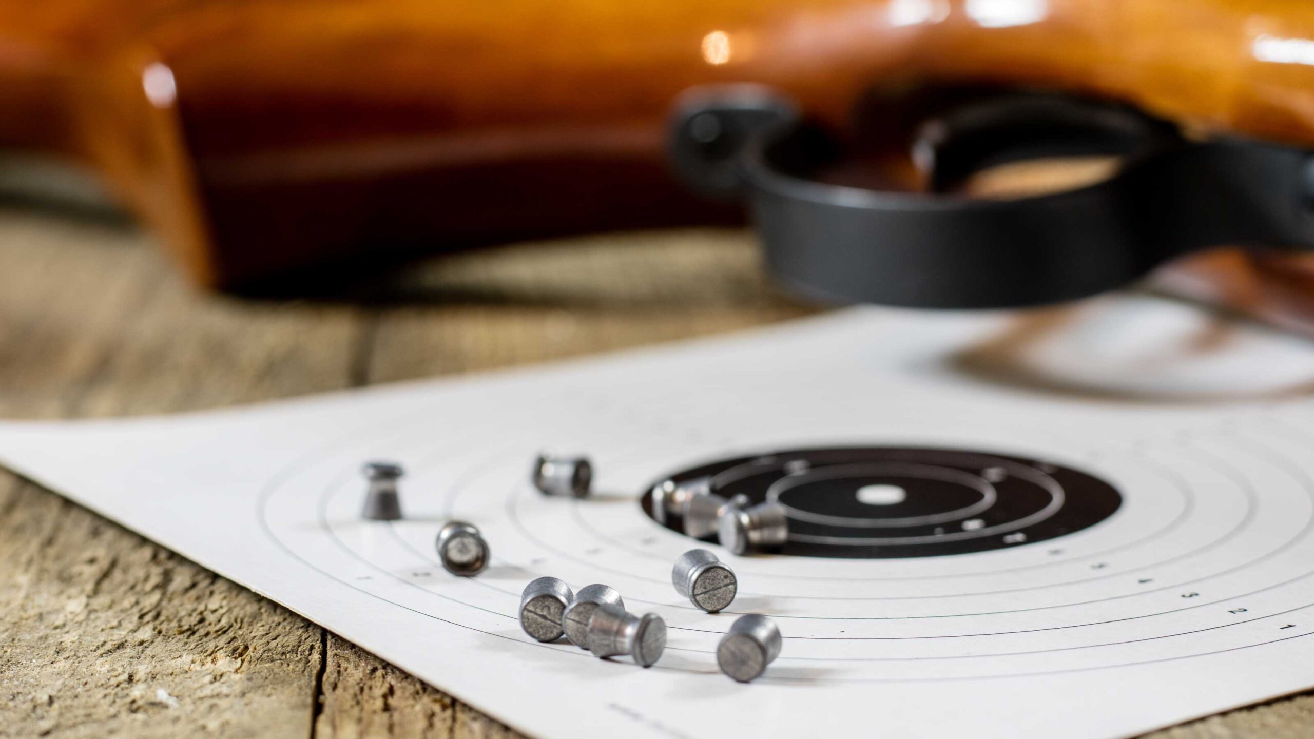 Close-up of air rifle pellets scattered on a paper shooting target with a rifle resting on a wooden table.