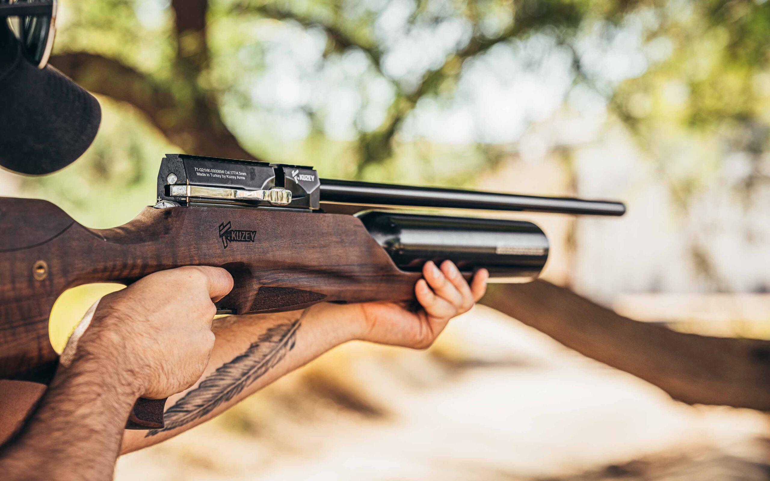 Person outdoors aiming a Kuzey air rifle with a wooden stock and mounted scope, wearing sunglasses and a cap, with a feather tattoo visible on the forearm and trees blurred in the background